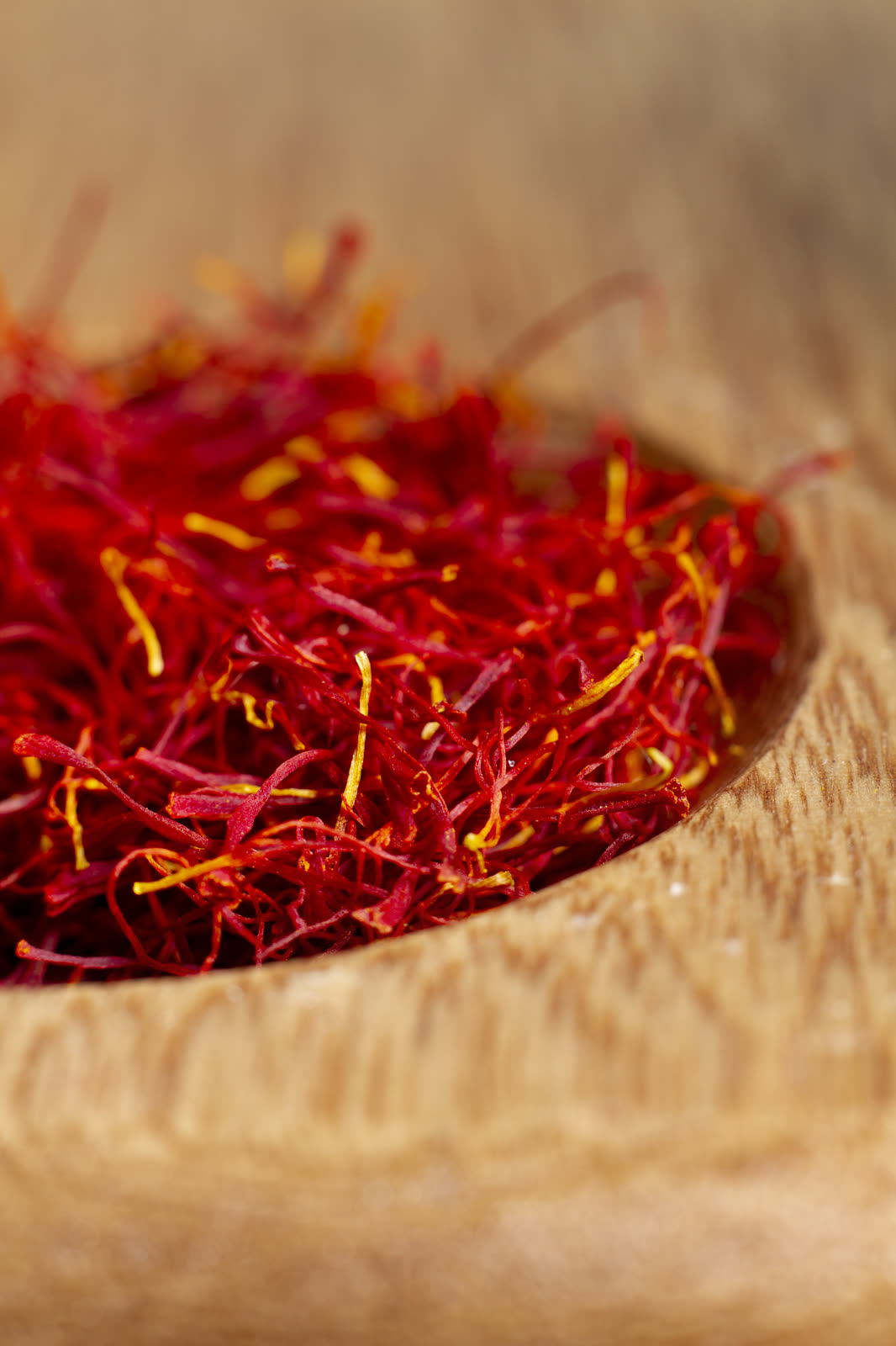 A heap of crimson saffron threads with a few golden-yellow styles, piled in a pale wooden bowl.